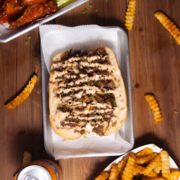 Cheesesteak sandwich with sauce, crinkle-cut fries, and wings on a wooden table at Sauce Wing Co.
