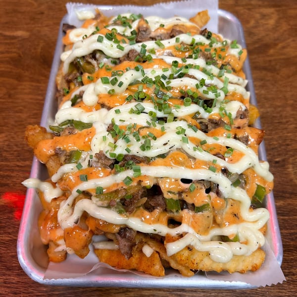 Loaded fries with steak, green peppers, orange sauce, white sauce, and chives in a metal tray.